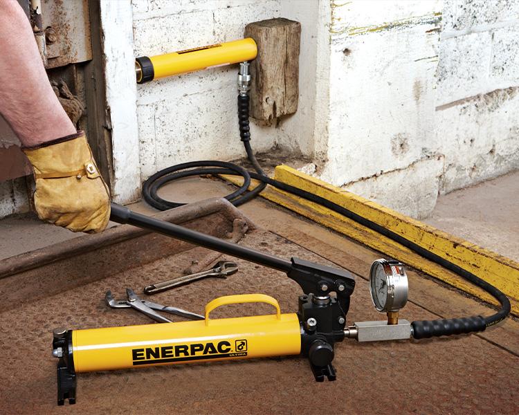 Workers using an enerpac handpump to manually extend a cylinder placed between two beams.