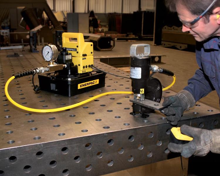 Construction worker using a punch tool connected with hydraulic hoses to an enerpac branded pump on a metal work desk.